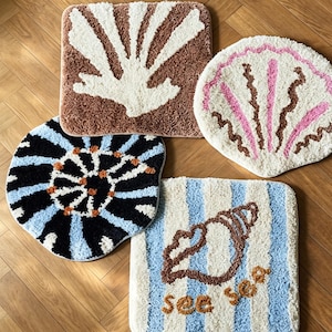 Puede incluir: Cuatro alfombras de mechones con diseños distintos: un cuadrado marrón con un motivo de coral blanco, una forma de concha blanca con detalles en rosa y marrón, una alfombra circular negra y azul claro con puntos naranjas y un cuadrado a rayas.