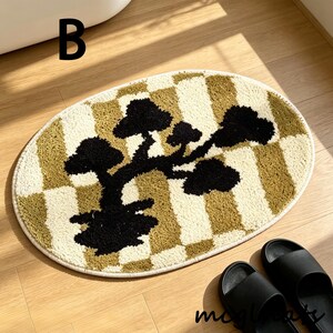 May include: Oval-shaped bath mat with a bonsai tree design. The mat features a checkered pattern in beige and olive green, with a black tree silhouette. The mat is placed on a wooden floor, with black sandals nearby.