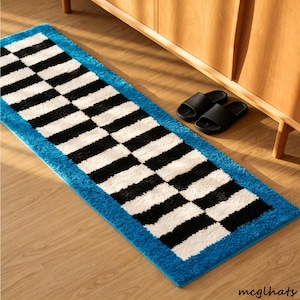 May include: A rectangular runner rug featuring a repeating black and white checkered pattern arranged in diagonal rows, bordered by a solid bright blue textured frame. The rug is placed on a light wood floor next to a pair of black slide sandals.