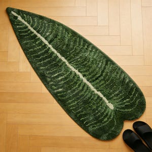 May include: A green leaf-shaped rug with a white central vein. The rug has a textured surface and is placed on a wooden floor. Black sandals are visible in the bottom right corner.
