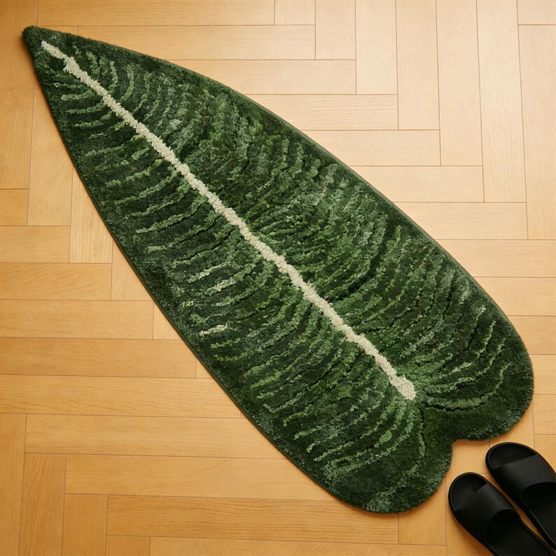 May include: A green leaf-shaped rug with a white central vein. The rug has a textured surface and is placed on a wooden floor. Black sandals are visible in the bottom right corner.