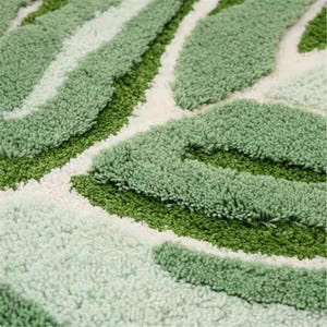 May include: Close-up of a green and white leaf-shaped rug. The rug has a textured, tufted design with varying shades of green and white, creating a natural, organic pattern. The rug is likely made of a soft, plush material.