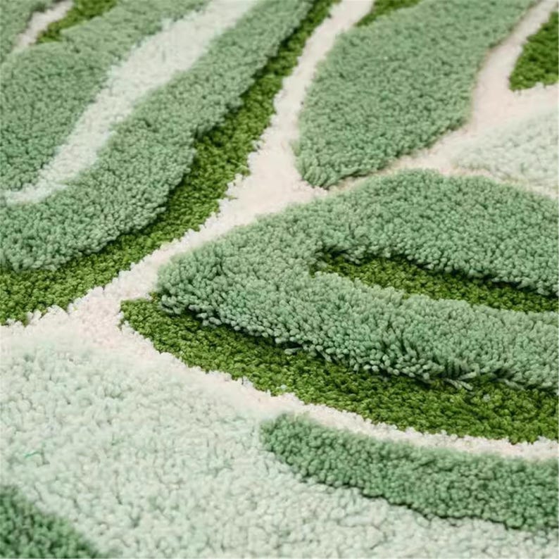 May include: Close-up of a green and white leaf-shaped rug. The rug has a textured, tufted design with varying shades of green and white, creating a natural, organic pattern. The rug is likely made of a soft, plush material.