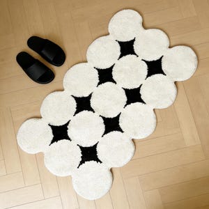 Op de afbeelding: Een crèmekleurig, getuft tapijt met een uniek ontwerp van onderling verbonden cirkels en zwarte vierkante accenten. Het tapijt wordt gecombineerd met twee zwarte sandalen, wat een moderne en stijlvolle uitstraling creëert.