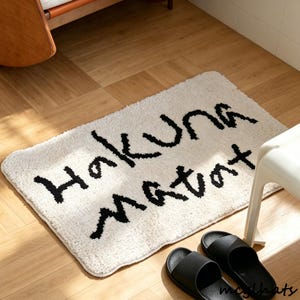 Può includere: Tappetino da bagno rettangolare color crema con le parole "Hakuna Matata" in corsivo nero. Due sandali neri sono posizionati vicino al tappetino. Il tappetino è su un pavimento in legno.