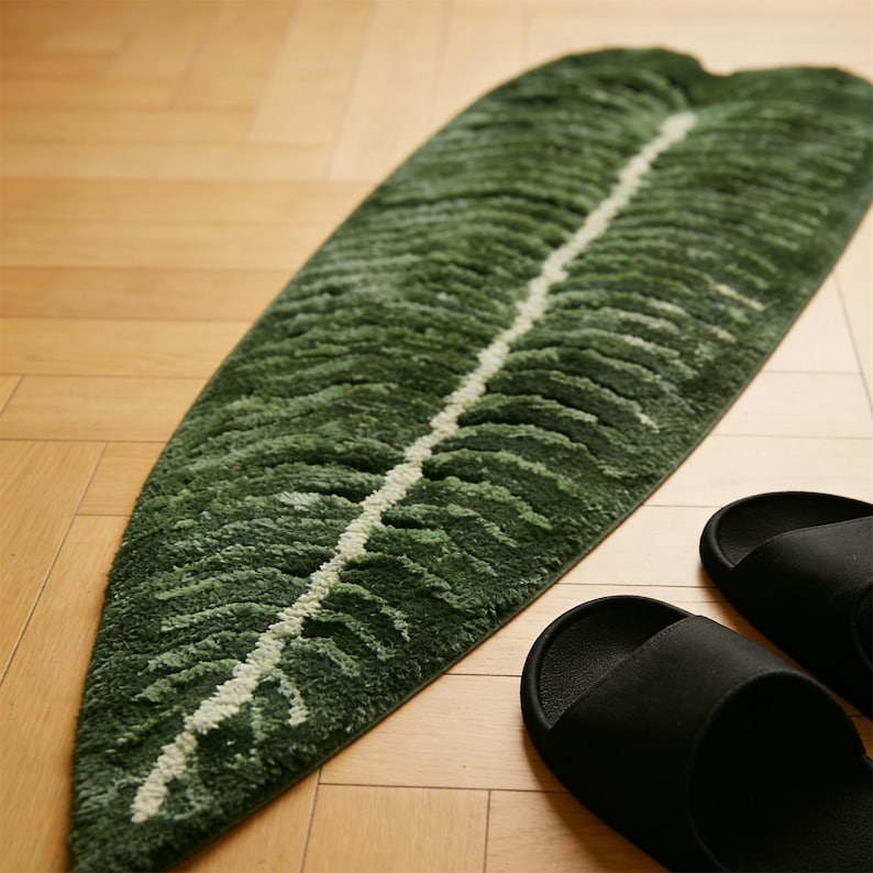 May include: A green leaf-shaped rug with a white central vein, placed on a wooden floor. The rug's textured surface features varying shades of green. Black sandals are visible near the rug. The rug is approximately 60 inches long.