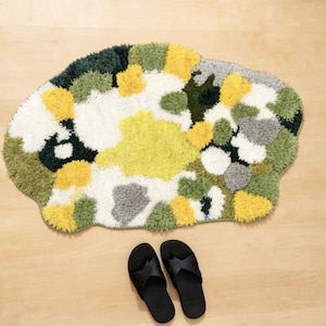 May include: An abstract rug featuring a mix of yellow, green, grey, and white tufted patterns. Two black flip-flops are placed below the rug on a light wood floor. The rug has an irregular, cloud-like shape.