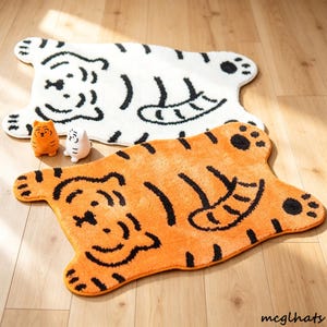 Könnte beinhalten: Zwei tigerförmige Teppiche, einer weiß mit schwarzen Streifen und einer orange mit schwarzen Streifen, jeweils mit einem Cartoon-Tigergesicht, einem geringelten Schwanz und Pfotenabdrücken. Zwei kleine Tigerfiguren stehen zwischen den Teppichen.