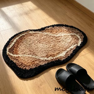 May include: A brown, beige, and black rug with an organic shape. The rug has a black border, a beige inner border, and a brown center. Two black sandals are next to the rug on a wooden floor.