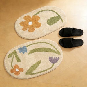Può includere: Due tappeti da bagno ovali beige con disegni floreali arancioni, blu e viola. Due pantofole nere sono accanto ai tappeti. I tappeti hanno foglie e steli verdi. Lo sfondo è un pavimento in legno marrone chiaro.