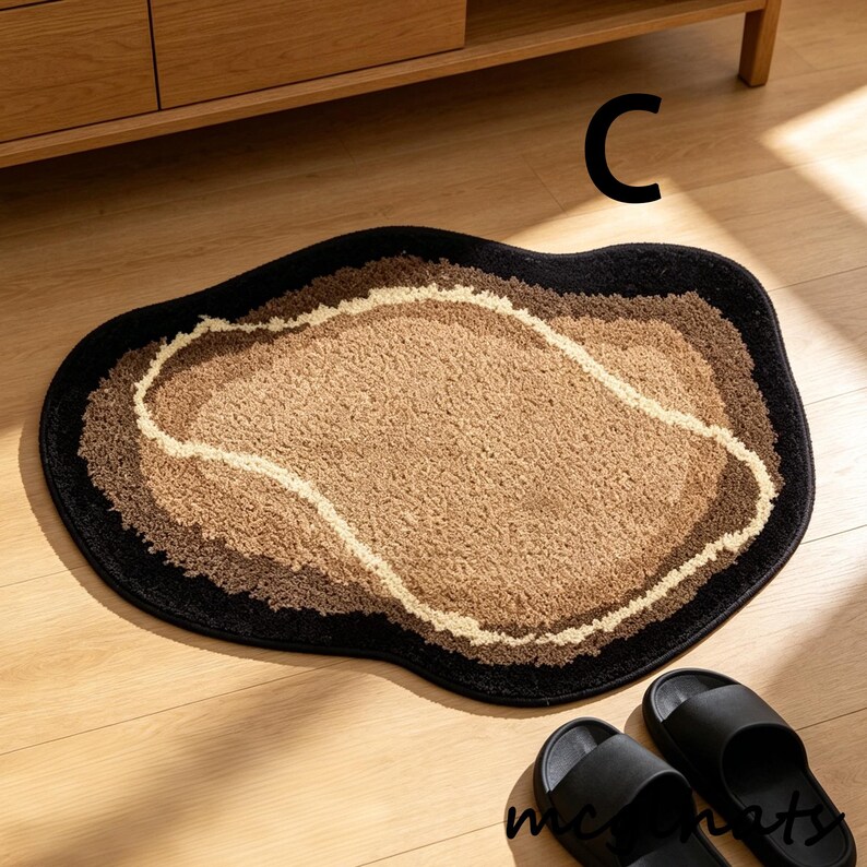 May include: A brown and black bath mat with a unique, organic shape. The mat features a layered design with a light brown center, surrounded by darker brown and black borders. The letter "C" is visible on the floor.