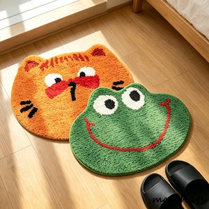 Könnte beinhalten: Zwei Cartoon-Teppiche: eine orangefarbene Katze mit roten Akzenten und ein grüner Frosch mit rotem Lächeln. Die Teppiche bestehen aus einem weichen, strukturierten Material und liegen auf einem Holzboden. Schwarze Sandalen sind im Vordergrund zu sehen.