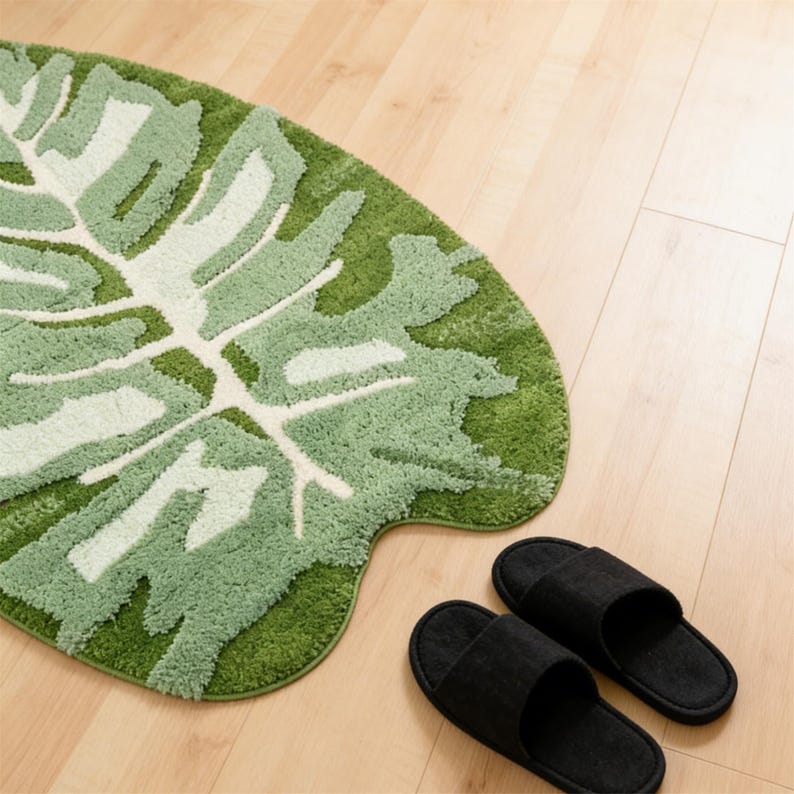 May include: A green leaf-shaped bath mat with a white and light green pattern, alongside a pair of black slippers. The bath mat is made of a soft, textured material, and the slippers have a simple, minimalist design.