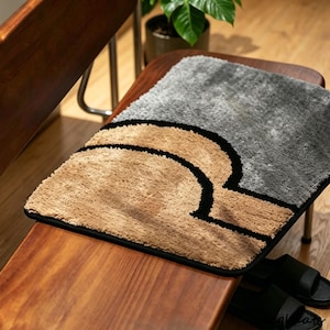 May include: A rectangular, plush, shaggy rug featuring a two-tone design. The top section is grey, while the bottom section is tan, separated by a bold, curved black line that forms a semi-circular shape. The rug has a black fabric border.