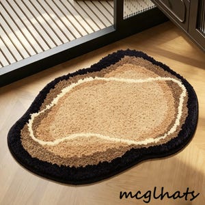 May include: A plush, irregularly shaped rug with a layered design. The rug features a black outer border, followed by shades of brown and beige, creating a textured, inviting surface. The rug is placed on a wooden floor.