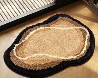 May include: A plush, irregularly shaped rug with a layered design. The rug features a black outer border, followed by shades of brown and beige, creating a textured, inviting surface. The rug is placed on a wooden floor.