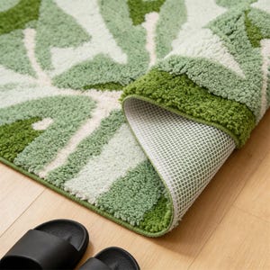May include: A rolled-up bath mat with a leaf pattern in shades of green and white. The mat has a non-slip backing. The rolled edge reveals the white backing. Black sandals are on a wooden floor.