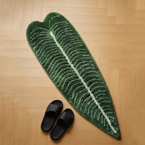 May include: A green leaf-shaped bath mat with a white central vein and textured details. Two black slide sandals are placed near the mat on a light wood floor. The mat is approximately 60 inches long.