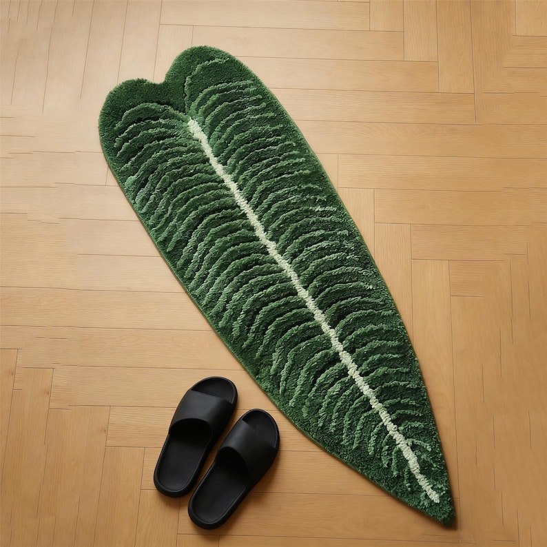 May include: A green leaf-shaped bath mat with a white central vein and textured details. Two black slide sandals are placed near the mat on a light wood floor. The mat is approximately 60 inches long.