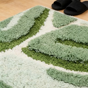 May include: A green and white patterned bath mat with a textured, plush surface. The mat features a geometric design with varying shades of green and white. Black slippers are visible in the background, resting on a wooden floor.