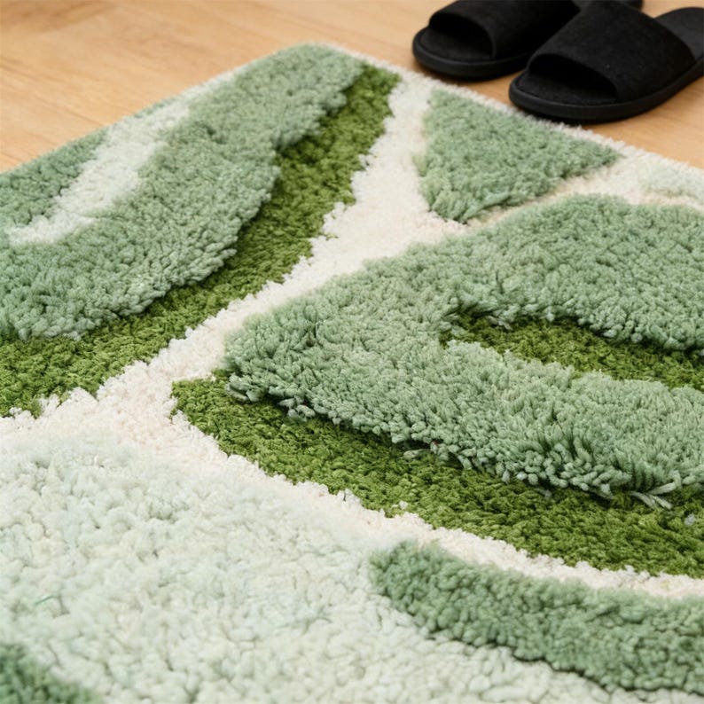 May include: A green and white patterned bath mat with a textured, plush surface. The mat features a geometric design with varying shades of green and white. Black slippers are visible in the background, resting on a wooden floor.