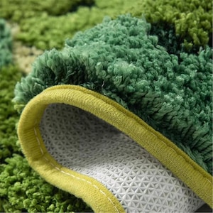 May include: Close-up of a green and yellow rug. The rug has a plush, textured surface with varying shades of green. The edge is trimmed with a yellow border and a white, patterned backing.