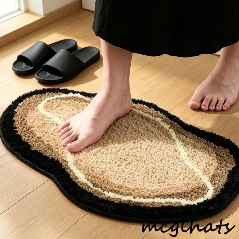 May include: A multi-toned oval bath mat with a black border, a light beige center, and a white accent. Black slide sandals are visible in the background. The mat is on a wooden floor.
