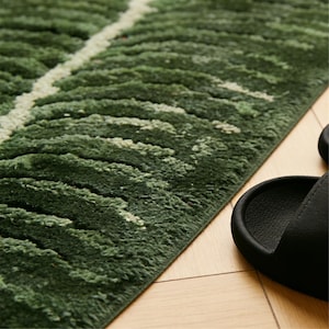 May include: Close-up of a green rug with a leaf-like design, featuring varying shades of green and a white stripe. The rug has a textured, plush appearance. A black sandal is partially visible on a wooden floor.