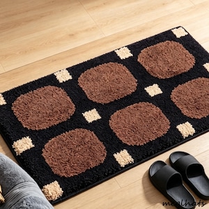 May include: A rectangular bath mat featuring a black base with a repeating pattern of large, rounded brown squares and small, cream-coloured square accents. The mat has a thick, textured pile surface and a dark border around the entire perimeter.