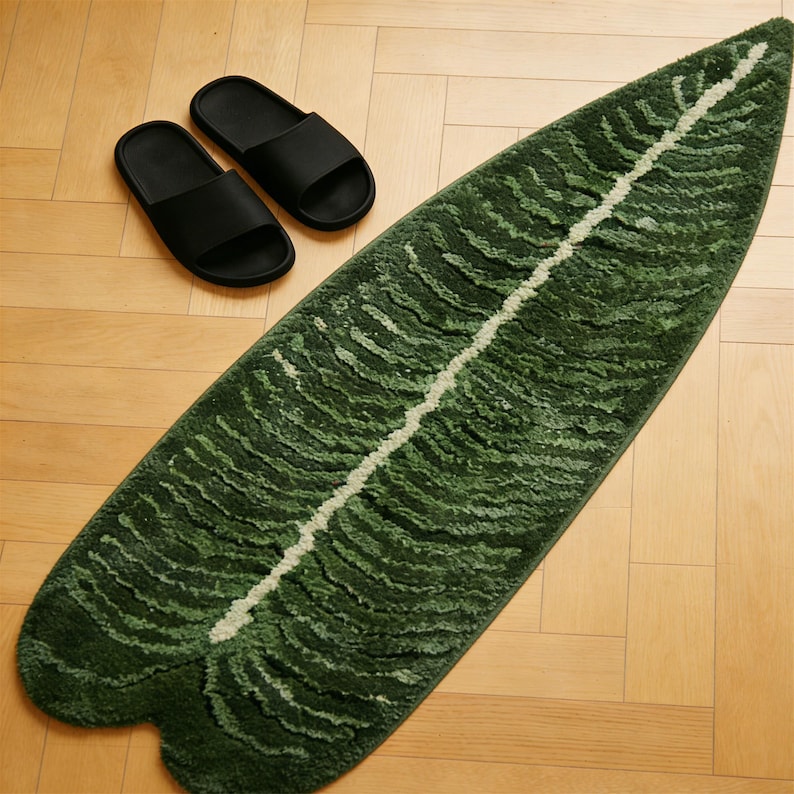 May include: A green leaf-shaped rug with a white central stripe and textured details, alongside two black slide sandals. The rug's design mimics a large leaf, adding a natural element to the space.