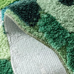 May include: Close-up of a green and white bath mat with a textured, shaggy surface. The mat features a geometric pattern of varying shades of green, with a white, non-slip backing visible. The edges are neatly finished with a sewn border.