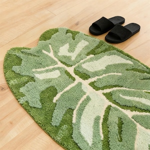 May include: A green and white leaf-shaped bath mat with a textured surface. The mat is made of a soft, plush material and features a detailed leaf design. Two black slippers are placed on the wooden floor next to the mat.