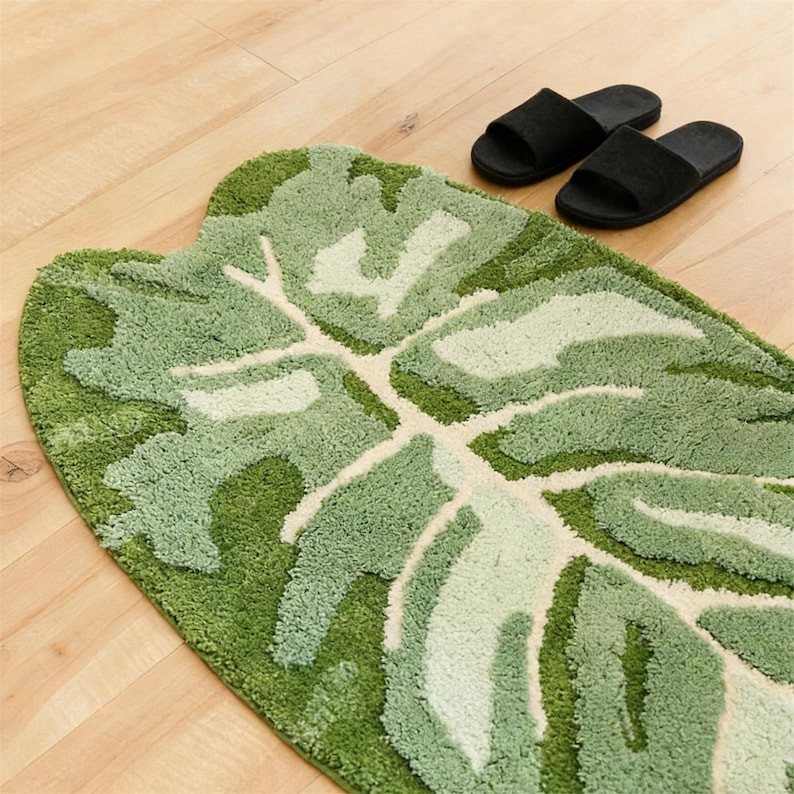 May include: A green and white leaf-shaped bath mat with a textured surface. The mat is made of a soft, plush material and features a detailed leaf design. Two black slippers are placed on the wooden floor next to the mat.