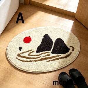 May include: An oval-shaped beige bath mat with a mountain and sun design. The mat features a red sun, two black mountain peaks, and brown water lines. The word "A" is printed in black on the top left of the mat.