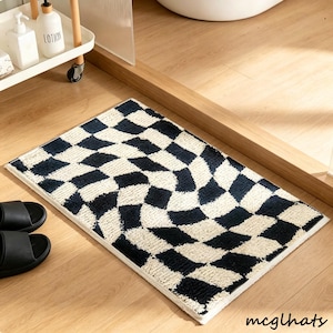 Puede incluir: Una alfombra de baño rectangular con un patrón de tablero de ajedrez en negro y crema. La alfombra está hecha de un material suave y texturizado y tiene un borde blanco. Se ven sandalias negras en el suelo de madera junto a la alfombra.