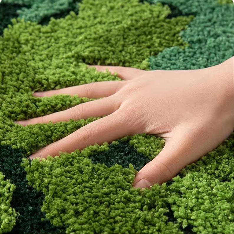 May include: A close-up shot of a green rug with a textured, shaggy surface. The rug features varying shades of green. A hand is pressing into the rug, showing its soft, plush texture. The rug appears to be made of a dense material.