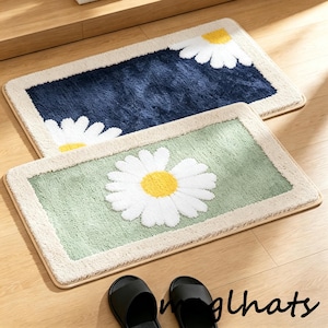 Op de afbeelding: Twee rechthoekige badmatten met madeliefjesontwerp. Eén mat is marineblauw met witte en gele madeliefjes, de andere is lichtgroen met een witte en gele madelief. Beide hebben een beige rand.