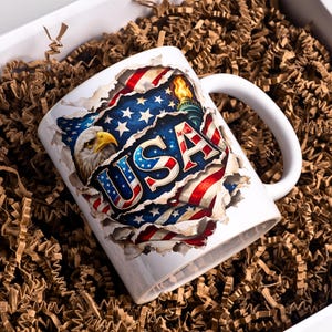 May include: White ceramic mug featuring a patriotic design with the word "USA" in red, white, and blue. The design includes an American flag, an eagle, and the Statue of Liberty. The mug is set in a box with brown packing material.