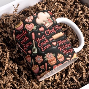 May include: A black ceramic mug with a white handle, decorated with pink and cream-colored baking-themed designs. The mug features text that reads "Made with Love", "Best Cook Ever", and "Mom's Kitchen Magic". The mug is nestled in brown paper filler.