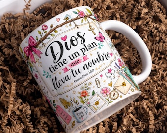 May include: A white ceramic mug featuring a colorful floral and butterfly illustration with the Spanish text 'Dios tiene un plan que lleva tu nombre' and 'Proverbios 16:9'. The design includes small icons like a cross, envelope, compass, and praying hands.