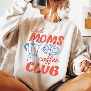 May include: A beige sweatshirt with the text "tired MOMS coffee CLUB" in red and blue. The graphic includes a coffee pot, croissant, and donut. The person is holding a black mug.