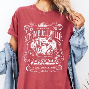 May include: A maroon t-shirt featuring a white graphic of Mickey Mouse from the 1928 Disney cartoon "Steamboat Willie." The design includes the text "Mickey Mouse" and "A Walt Disney Comic."