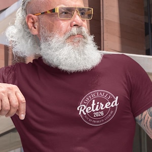 May include: Burgundy t-shirt with the text "Officially Retired 2026, Not My Problem Anymore" in a white circular design. The shirt is short-sleeved and made of a soft material. The person is wearing blue jeans.