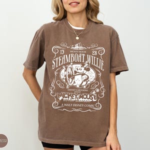 May include: Espresso-colored t-shirt featuring a vintage design with the text "Steamboat Willie" and "Mickey Mouse." The graphic includes an image of Mickey Mouse at the helm of a steamboat. The shirt is a relaxed fit.