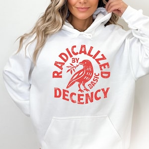 May include: A white hooded sweatshirt featuring a red graphic print on the chest. The design includes a bird holding a branch, surrounded by the text RADICALIZED BY BASIC DECENCY in a circular, distressed font, with a front kangaroo pocket.