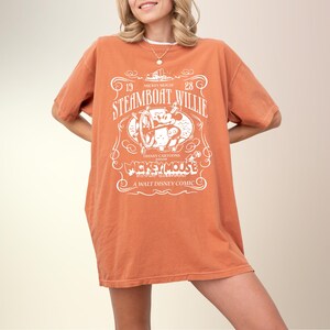 May include: An orange t-shirt featuring a vintage design of Mickey Mouse in Steamboat Willie. The white graphic includes the text "STEAMBOAT WILLIE", "MICKEY MOUSE", and "A WALT DISNEY COMIC". The shirt is a loose fit.
