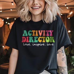 May include: A black short-sleeved t-shirt featuring the text "ACTIVITY DIRECTOR" in large, colorful, retro-style letters, with the words "love. inspire. care" printed in white underneath the main text in a simple, clean, cursive font.