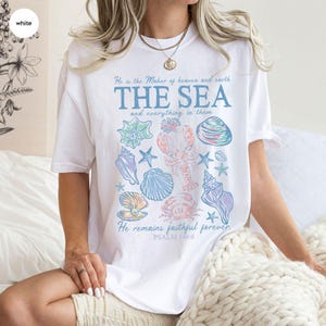 May include: A white short-sleeve t-shirt featuring a graphic print of various seashells, a lobster, and a crab. The text reads: He is the Maker of heaven and earth, THE SEA and everything in them, He remains faithful forever, PSALM 146:6.