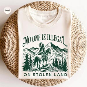 May include: Cream-colored t-shirt with a green graphic. The graphic depicts a Native American on horseback in front of mountains and trees, with the text "NO ONE IS ILLEGAL ON STOLEN LAND".