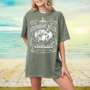 May include: An olive green t-shirt featuring a vintage design with the text "Steamboat Willie" and "Mickey Mouse". The graphic includes an illustration of Mickey Mouse and a steamboat. The shirt is a relaxed fit.
